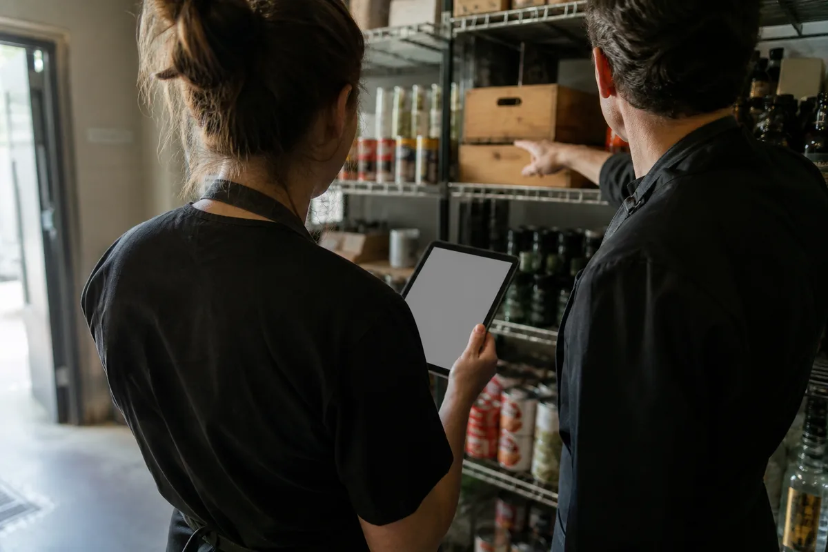 Deux membres d&rsquo;équipe en tablier notant des quantités sur une tablette devant une étagère de réserve